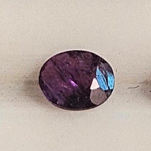 May include: Three oval-cut gemstones with a deep purple hue. The gems have facets that catch the light, displaying flashes of blue and white. The stones are set against a neutral background.