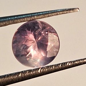 May include: A round, faceted gemstone held by tweezers. The gem has a blend of pink and purple hues, with light reflecting off its many facets. The tweezers are silver-colored and hold the gem against a soft, neutral background.