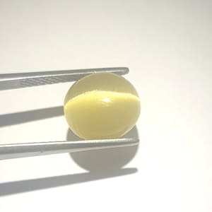 Puede incluir: Una gema redonda de color amarillo pálido sujeta con pinzas plateadas. La gema tiene una superficie lisa y pulida y un brillo interno sutil. La gema es una esfera.