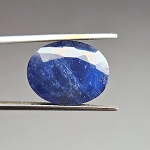 May include: A deep blue, oval-cut sapphire gemstone. The gem is held by tweezers, showcasing its facets and internal features. The sapphire's rich colour is contrasted against a neutral background, emphasizing its clarity and shape.