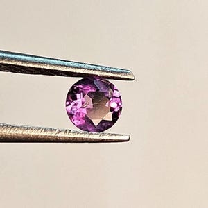 May include: A round, faceted gemstone held by tweezers. The gem is a vibrant purple color with a gradient effect, transitioning to a darker hue in the center. The tweezers are silver-colored.