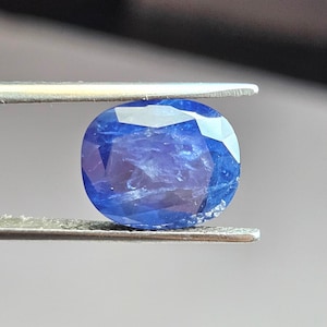 May include: A deep blue oval-shaped gemstone with a faceted cut. The gemstone is held by a pair of tweezers.