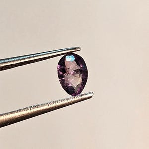 May include: A pear-shaped, faceted gemstone held by tweezers. The gem is a deep purple color with internal inclusions and a hint of blue iridescence. The tweezers are silver-colored and the background is a neutral tone.