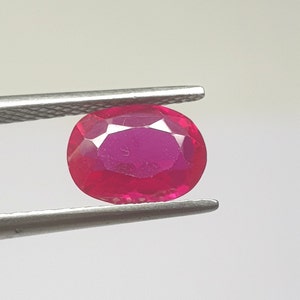 May include: A faceted, oval-cut ruby gemstone held by metal tweezers. The ruby is a deep, vibrant red color, with visible internal facets. The background is a plain, light gray.
