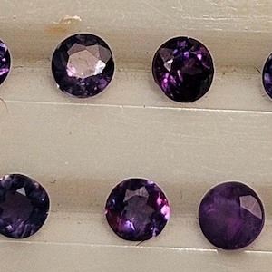 May include: Eight round, faceted gemstones in varying shades of purple and violet. The gems are arranged in two rows, each stone displaying a unique internal pattern and sparkle. The gems are set against a neutral background.