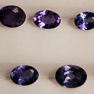 May include: Five oval and round-cut, deep purple gemstones are displayed. The gems have facets that catch the light, creating a sparkling effect. The stones vary slightly in shape and size, showcasing their natural beauty. The gemstones are arranged on a light-colored surface.