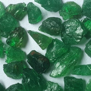May include: A collection of green gemstone roughs. The rough gemstones are various sizes and shapes. The gemstones are on a white background.