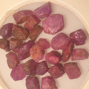 Puede incluir: Colección de piedras preciosas de rubí en bruto, en varios tonos de rojo y rosa. Las piedras, de forma irregular, presentan una textura natural y sin pulir. Los rubíes se muestran en un recipiente transparente y circular.
