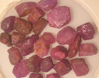 May include: A collection of rough-cut ruby gemstones in varying shades of red and pink. The gemstones are irregularly shaped and appear to be raw, with a natural, unpolished texture. The rubies are displayed in a clear, circular container.