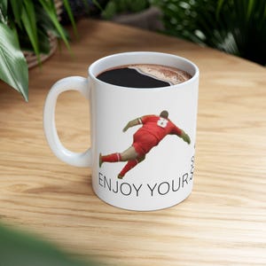 May include: White ceramic coffee mug with a black coffee fill. The mug features a graphic of a person in a red uniform, mid-action, with the text "ENJOY YOUR". The mug sits on a wooden surface.