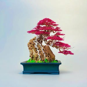 May include: A wire sculpture of a bonsai tree featuring a twisted metallic trunk and branches with vibrant pink foliage. The tree is anchored to a textured rock base with small green accents, all set within a rectangular dark blue ceramic pot.