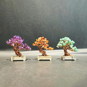 May include: Three miniature bonsai trees crafted with twisted copper wire trunks and branches. Each tree features small gemstone leaves in purple, orange, and light green, respectively, and is set in a rectangular off-white ceramic pot.