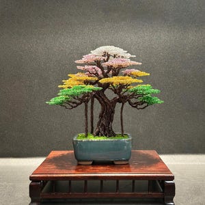 May include: A handcrafted bonsai tree with a multi-colored canopy of pink, white, yellow, and green. The tree sits in a teal rectangular pot on a dark wooden stand. The bonsai is a unique decorative item.