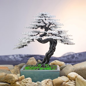 Op de afbeelding: Een bonsaiboomsculptuur van draad met een gedraaide zwarte metalen stam en takken versierd met heldere, kristallijne kralen die bladeren voorstellen. De boom staat in een rechthoekige blauwgroene keramische pot gevuld met groen mos.