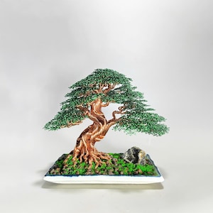 May include: A detailed bonsai tree sculpture. The tree has a copper wire trunk and branches with green wire leaves. It is set on a rectangular base with moss and a small rock. The bonsai is a miniature representation of a mature tree.