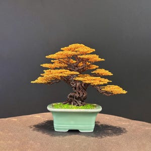 May include: A handcrafted bonsai tree sculpture with a twisted brown trunk and vibrant orange-yellow foliage. The miniature tree sits in a light green rectangular ceramic pot with a bed of green moss. The background is a dark gray.
