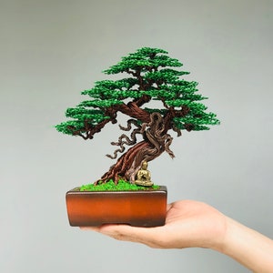 May include: A wire sculpture of a bonsai tree with a green canopy and a brown trunk. The tree is in a brown pot and a small gold figurine is sitting at the base of the trunk.