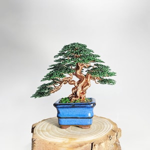 May include: A handcrafted bonsai tree sculpture with a copper wire trunk and green beaded foliage. The tree is planted in a square, blue ceramic pot and sits on a wooden base. The bonsai is a miniature representation of a mature tree.