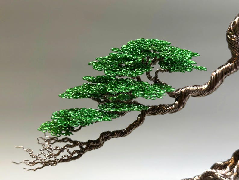 May include: A wire sculpture of a bonsai tree with a twisted trunk and green leaves. The tree is made of copper wire and green beads.