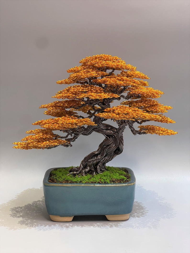 Puede incluir: Una escultura de alambre de un &aacute;rbol bons&aacute;i con un tronco marr&oacute;n y hojas naranjas. El &aacute;rbol est&aacute; en una maceta de cer&aacute;mica azul con una base de musgo verde.