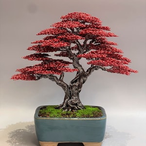 May include: A red wire bonsai tree sculpture in a blue ceramic pot with green moss. The tree has a twisted trunk and branches with red leaves.