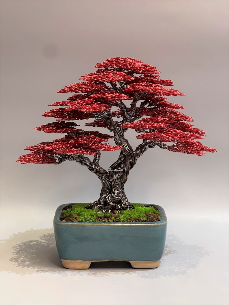 May include: A red wire bonsai tree sculpture in a blue ceramic pot with green moss. The tree has a twisted trunk and branches with red leaves.