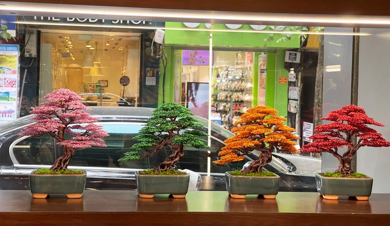 May include: Four bonsai trees in different colored beads, pink, green, orange, and red, in small green pots on a wooden surface.