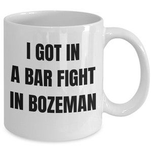 May include: White ceramic coffee mug with black text that reads "I got in a bar fight in Bozeman".