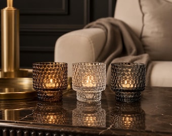 May include: Three cylindrical glass candle holders with a textured honeycomb pattern. The set includes one amber-colored, one clear, and one dark gray glass holder, each containing a small lit candle that casts a warm glow through the glass.