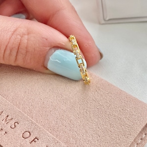 May include: A gold chain ring with small clear stones set into the links. The ring is on a pink suede surface with the text "MS OF BELLI" visible.