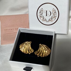 May include: A pair of gold-toned earrings in the shape of a fan, displayed in a white box with a black interior. The box has a logo with the letter "D" surrounded by a wreath of leaves.