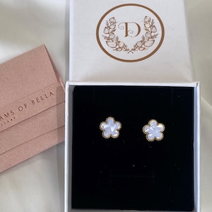 May include: A pair of white flower-shaped earrings with gold accents in a white box with a gold logo that says "D" inside a wreath. The box is lined with black velvet.