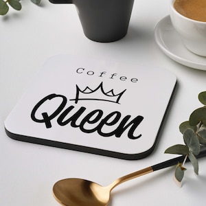 Könnte beinhalten: Ein quadratischer weißer Untersetzer mit schwarzem Rand zeigt die Worte "Coffee Queen" in einer Schreibschrift mit einer Kronengrafik. Ein goldener Löffel und zwei Kaffeetassen sind ebenfalls zu sehen.