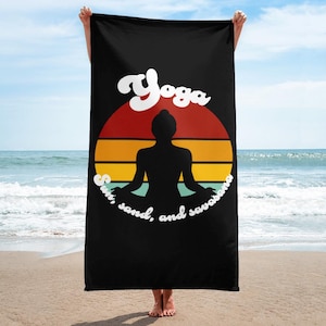 Op de afbeelding: Zwarte strandhanddoek met een retro zonsondergangontwerp en een silhouet van een persoon die yoga doet. De tekst "Yoga, zon, zand en savasana" is op de handdoek gedrukt.