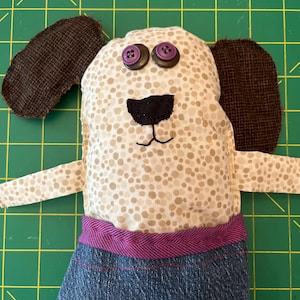Peut inclure: Une peluche en forme de chien avec un corps crème à pois marron, des oreilles en tissu texturé marron foncé et deux yeux en boutons violets. Le chien possède un nez en tissu noir, une bouche brodée et porte un pantalon en jean bleu à ceinture violette.