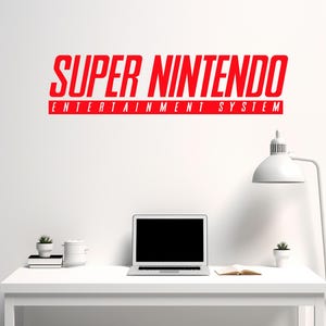 Peut inclure: Texte rouge "SUPER NINTENDO ENTERTAINMENT SYSTEM" sur un mur blanc au-dessus d'un bureau blanc. Un ordinateur portable, une petite plante en pot et un livre ouvert sont sur le bureau. Une lampe blanche est à droite.
