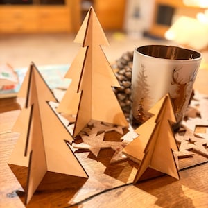 May include: Three wooden Christmas tree ornaments on a table with a star-shaped coaster. The trees are made of light brown wood and have a simple design.