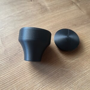 Op de afbeelding: Een zwarte flesstop en dop met een matte afwerking. De stop heeft een uitlopende, conische vorm, terwijl de dop een platte, cirkelvormige schijf is. Beide zijn gemaakt van een solide materiaal.