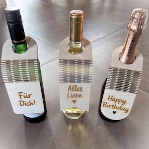 May include: Three bottles of wine with wooden gift tags. The tags have laser-cut designs and text that reads "Für Dich!", "Alles Liebe", and "Happy Birthday". The bottles are various colors, including green, gold, and rose.