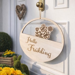 May include: A wooden circular door sign with the words "Hallo Frühling" and a floral design. The sign is hanging on a white door with a gold doorknob. A woven heart-shaped decoration is visible.