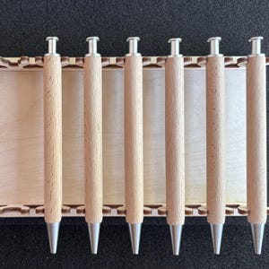 Könnte beinhalten: Ein Set aus sechs Holzstiften mit silberfarbenen Spitzen und Oberteilen, angeordnet in einem hellen Holzhalter. Die Stifte sind zylindrisch und haben eine natürliche Holzmaserung. Der Halter hat ein dekoratives, lasergeschnittenes Design.