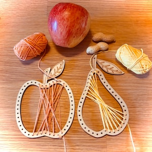 Può includere: Due kit di artigianato in legno a forma di mela e pera. Il kit della mela è in fase di infilaggio con filo arancione e il kit della pera con filo giallo. Sullo sfondo ci sono due gomitoli di lana e una mela rossa.
