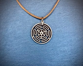 Puede incluir: Un collar de madera con un cordón de cuero marrón. El colgante es de diseño circular con una estrella de cinco puntas en el centro y círculos concéntricos que irradian hacia afuera.