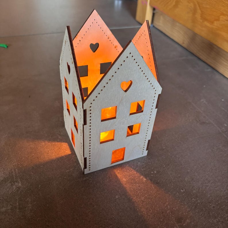 May include: A wooden house-shaped candle holder with a warm orange glow emanating from within. The white exterior features heart-shaped cutouts and square windows. The edges are a darker brown, and the candle holder is on a gray surface.