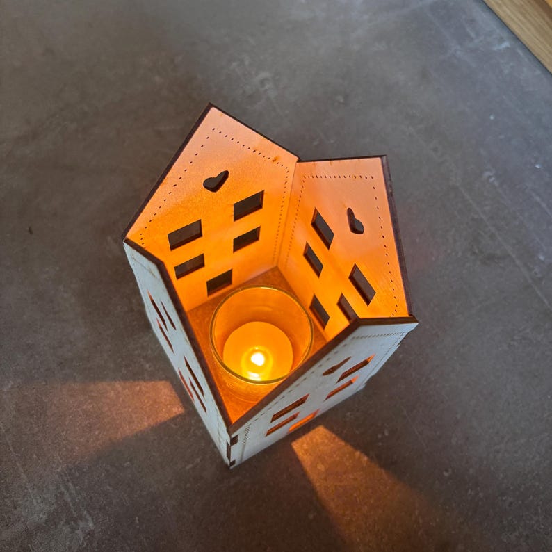 May include: A wooden house-shaped candle holder with rectangular and heart-shaped cutouts. A lit candle in a glass holder sits inside, casting a warm glow. The candle holder is made of light-colored wood with dark brown edges.