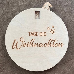 Può includere: Ornamento rotondo natalizio in legno con il numero 24 visibile in una piccola finestra. Il testo "TAGE BIS Weihnachten" è inciso sulla superficie, insieme a stelle e un cappello di Babbo Natale. L'ornamento è in legno chiaro.