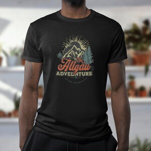 Può includere: Maglietta nera con una grafica in stile vintage di montagne, un sole e alberi. Il testo "The Allgäu Adventure" è stampato in un font retrò con le parole "Explore the Mountain" sotto.