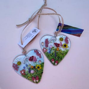 May include: Two heart-shaped fused glass ornaments featuring colourful floral meadow designs with bees and mushrooms. Each piece includes a tan ribbon for hanging and a small white tag with text. The glass displays vibrant green, yellow, red, and pink accents.