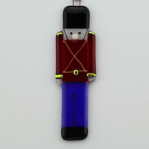 May include: A glass ornament shaped like a toy soldier. The soldier is wearing a red uniform with gold trim, blue pants, and a black hat.