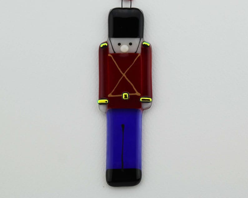 May include: A glass ornament shaped like a toy soldier. The soldier is wearing a red uniform with gold trim, blue pants, and a black hat.